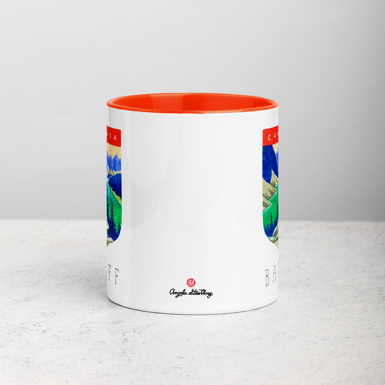 White ceramic coffee mug with orange handle and inside; has Banff National Park illustration by Angela Staehling