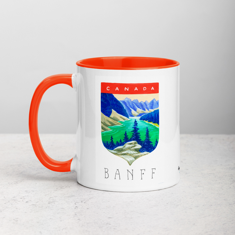 White ceramic coffee mug with orange handle and inside; has Banff National Park illustration by Angela Staehling