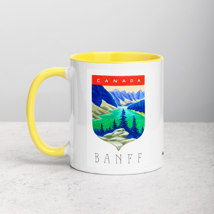 White ceramic coffee mug with yellow handle and inside; has Banff National Park illustration by Angela Staehling