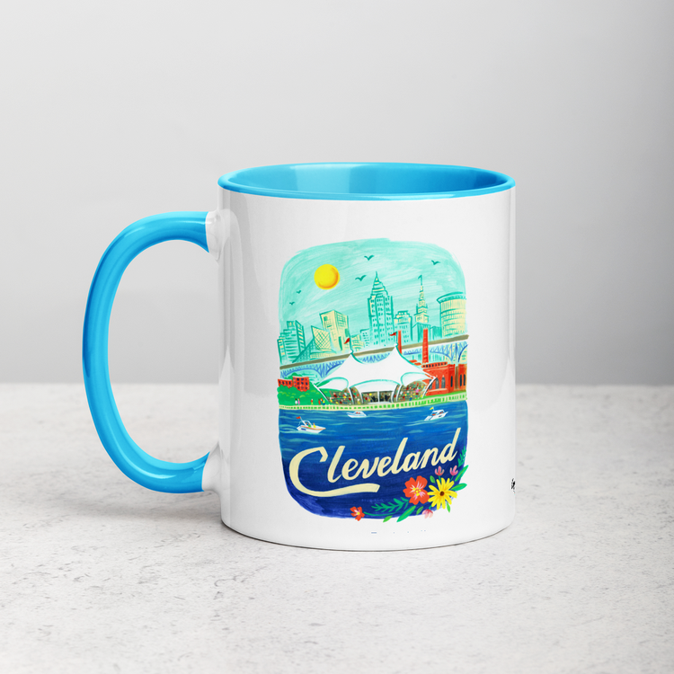 White ceramic coffee mug with blue handle and inside; has Cleveland Ohio illustration by Angela Staehling