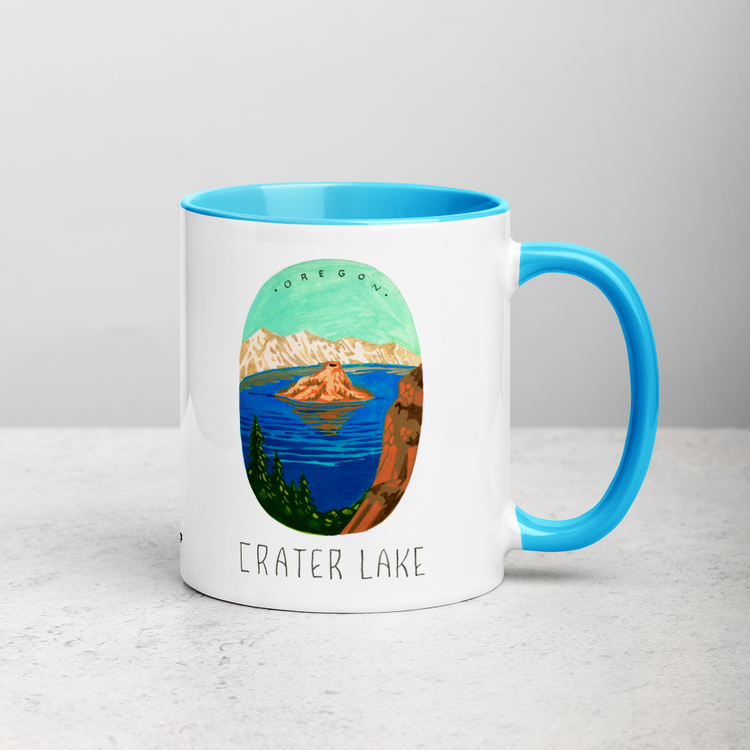 White ceramic coffee mug with blue handle and inside; has Crater Lake National Park illustration by Angela Staehling