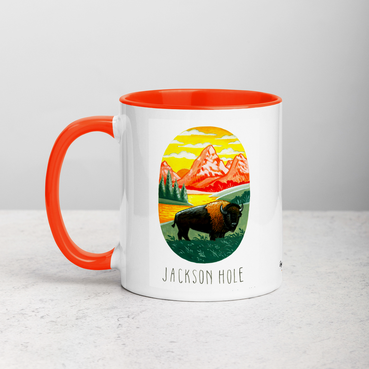 White ceramic coffee mug with orange handle and inside; has Jackson Hole National Park illustration by Angela Staehling