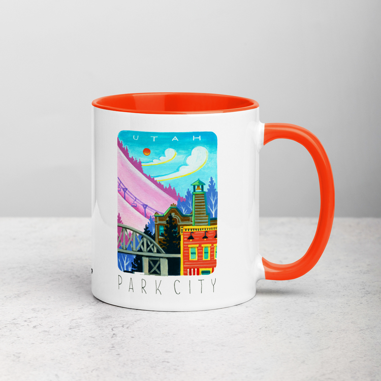 White ceramic coffee mug with orange handle and inside; has Park City illustration by Angela Staehling