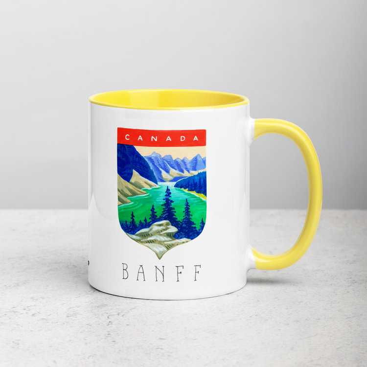 White ceramic coffee mug with yellow handle and inside; has Banff National Park illustration by Angela Staehling