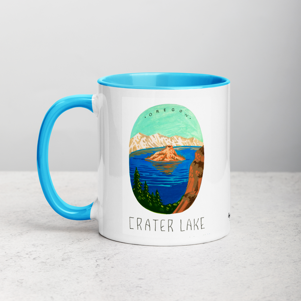 White ceramic coffee mug with blue handle and inside; has Crater Lake National Park illustration by Angela Staehling
