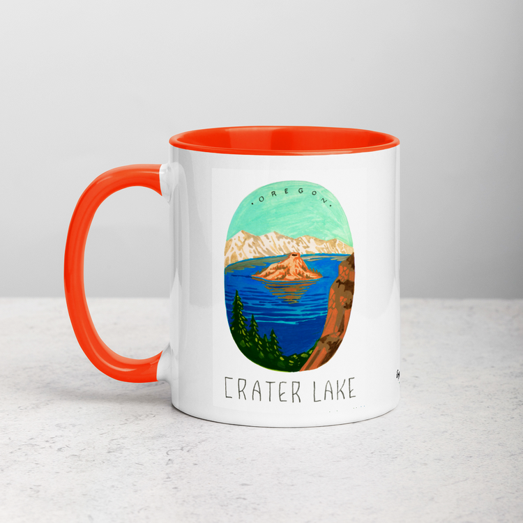 White ceramic coffee mug with orange handle and inside; has Crater Lake National Park illustration by Angela Staehling
