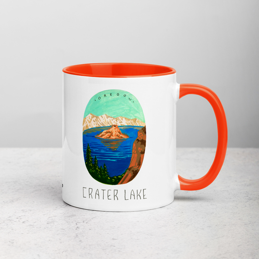 White ceramic coffee mug with orange handle and inside; has Crater Lake National Park illustration by Angela Staehling