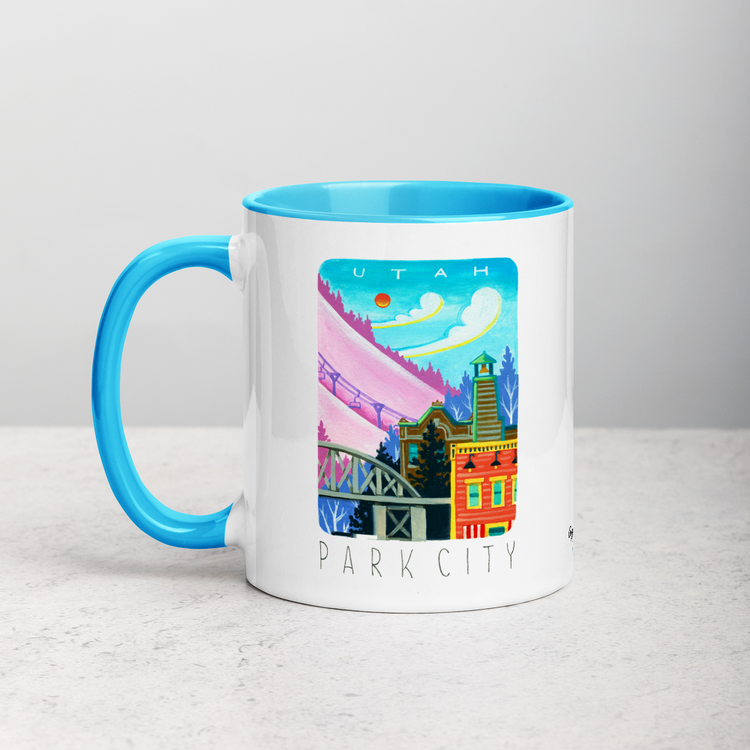 White ceramic coffee mug with blue handle and inside; has Park City illustration by Angela Staehling