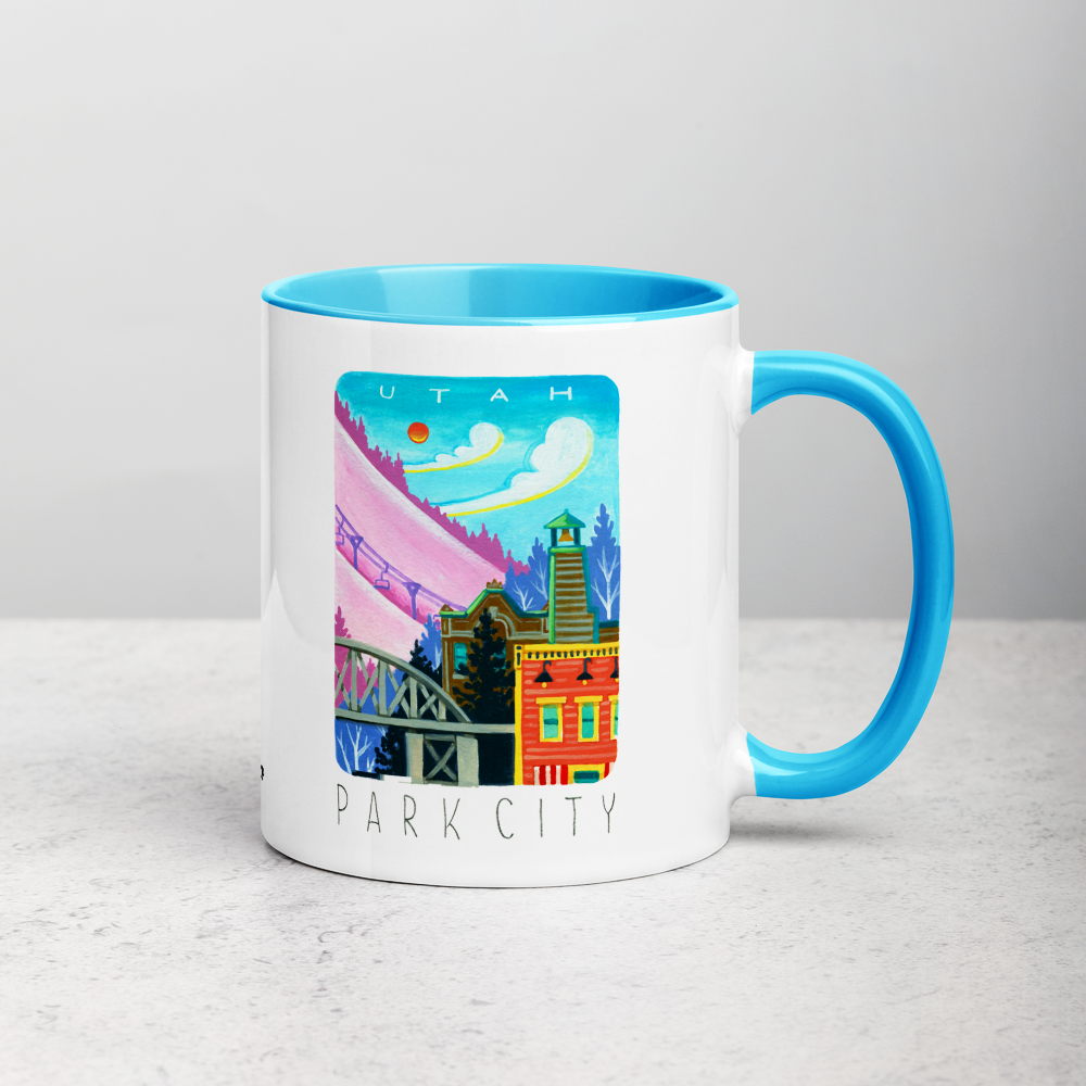 White ceramic coffee mug with blue handle and inside; has Park City illustration by Angela Staehling