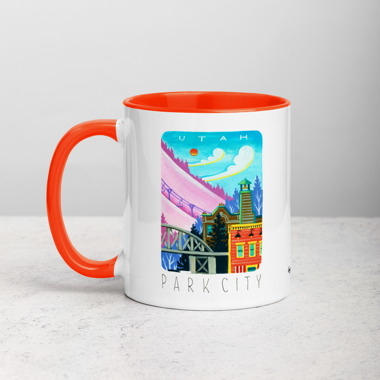 White ceramic coffee mug with orange handle and inside; has Park City illustration by Angela Staehling