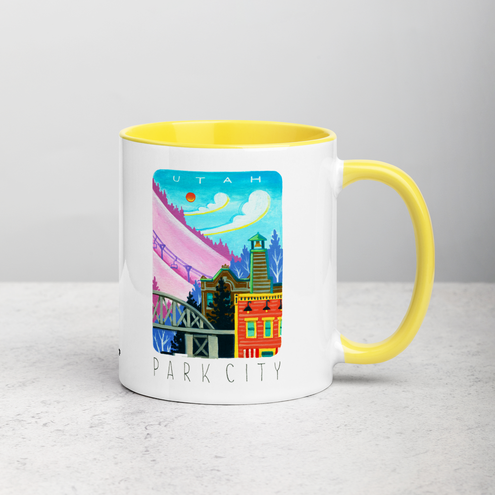 White ceramic coffee mug with yellow handle and inside; has Park City illustration by Angela Staehling