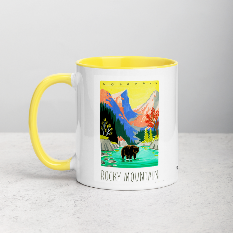 White ceramic coffee mug with yellow handle and inside; has Rocky Mountain National Park illustration by Angela Staehling