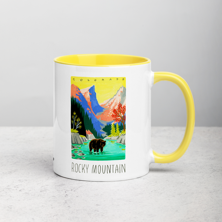 White ceramic coffee mug with yellow handle and inside; has Rocky Mountain National Park illustration by Angela Staehling